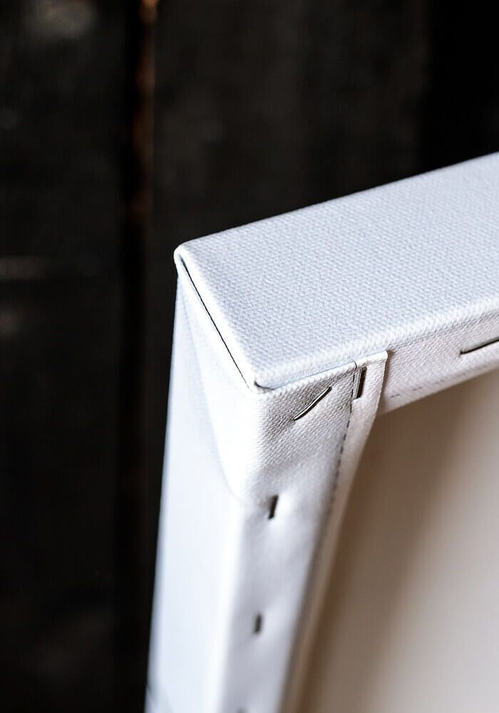 Close up of a white canvas print or framed wall art corner, showing the textured fabric surface and edge construction against a dark blurred background.