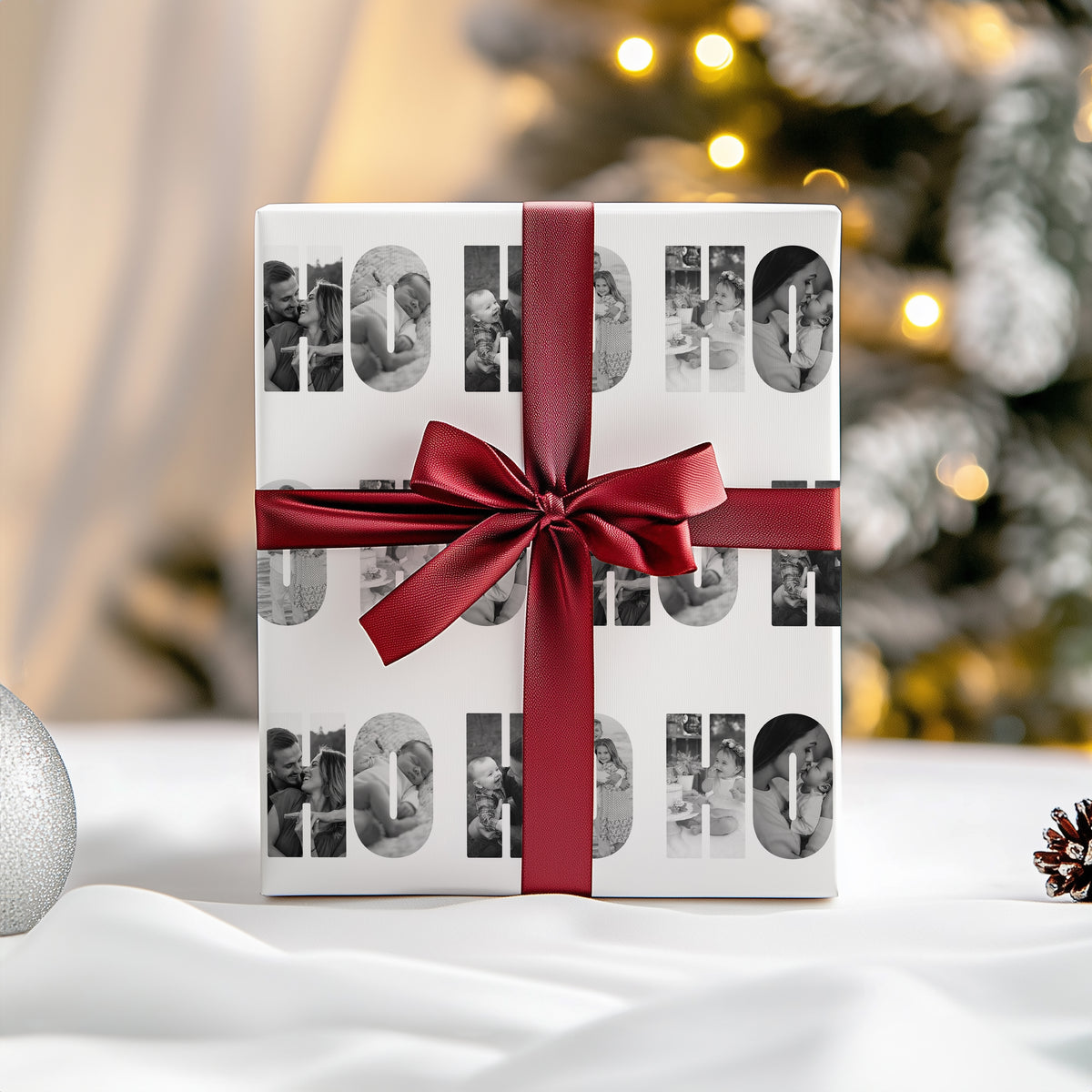 A square gift box wrapped in white paper with large black-and-white photo collage letters spelling 'HO HO HO' and tied with a dark red satin ribbon and bow, set on a white surface with a softly blurred Christmas tree in the background.