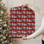 A person in a cream cable knit sweater holds up a large red gift wrap with a repeating pattern of black and white holiday photos within the text 'HO HO HO'. A decorated Christmas tree with warm lights is visible in the background