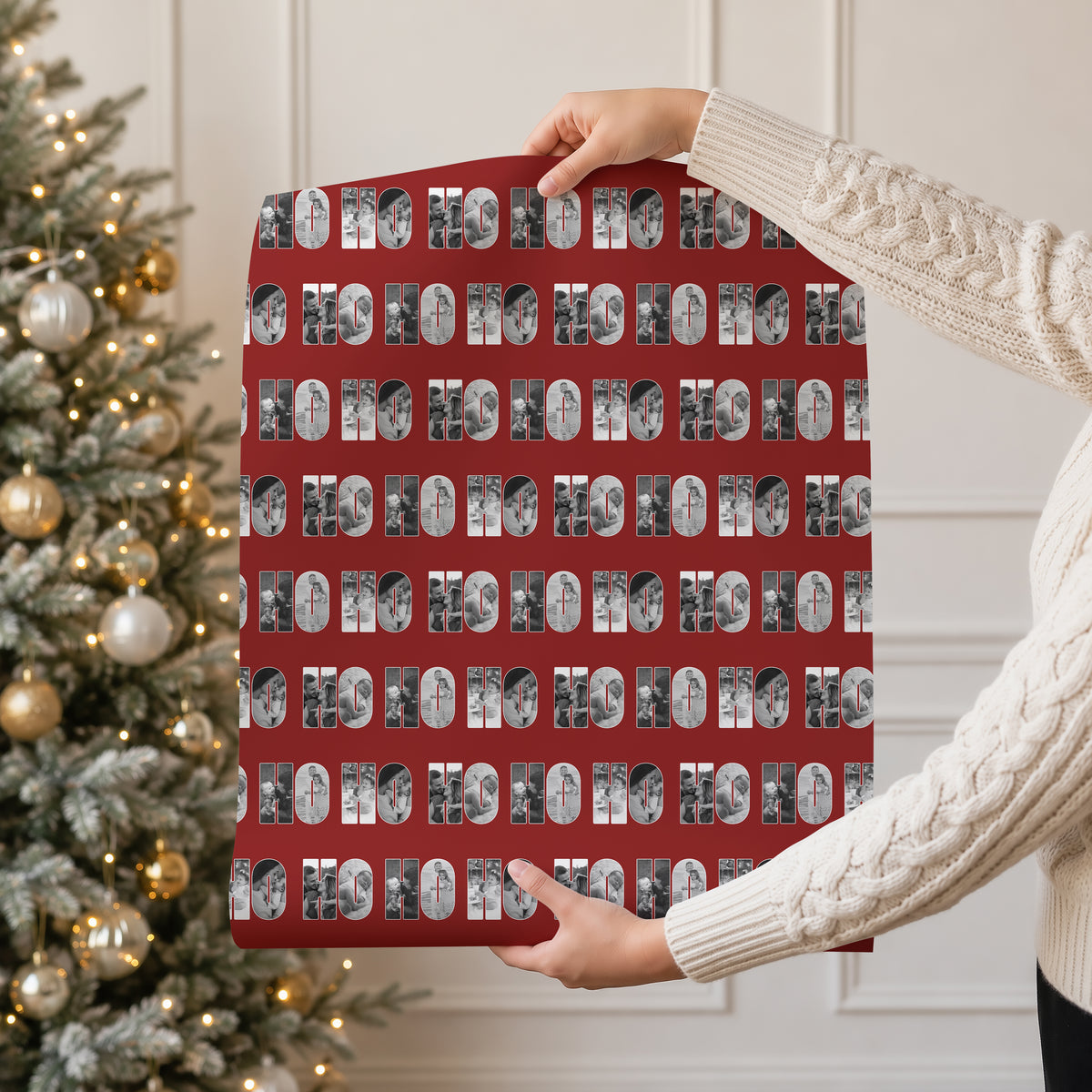 A person in a cream cable knit sweater holds up a large red gift wrap with a repeating pattern of black and white holiday photos within the text 'HO HO HO'. A decorated Christmas tree with warm lights is visible in the background