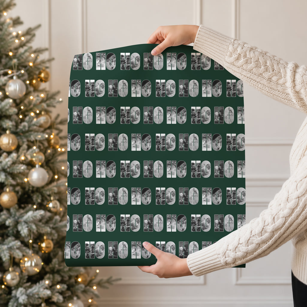 A person in a cream cable knit sweater holds up a large rectangular sheet of wrapping paper featuring a repeating pattern of black and white photos within the text 'HO HO HO
 on a dark green background. A decorated Christmas tree with warm lights is visible in the background.