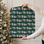 A person in a cream cable knit sweater holds up a large rectangular sheet of wrapping paper featuring a repeating pattern of black and white photos within the text 'HO HO HO
 on a dark green background. A decorated Christmas tree with warm lights is visible in the background.