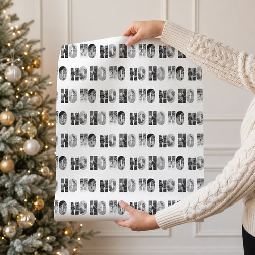 A person in a cream cable knit sweater holds up a roll of wrapping paper featuring a repeating black and white photos within 'HO HO HO' pattern, with a decorated Christmas tree in the background.