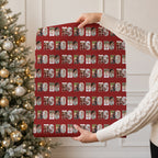 A person in a cream cable knit sweater holds up a large sheet of wrapping paper featuring a repeating Christmas themed pattern. The pattern consists of a red background with photos within the text 'HO HO HO'.