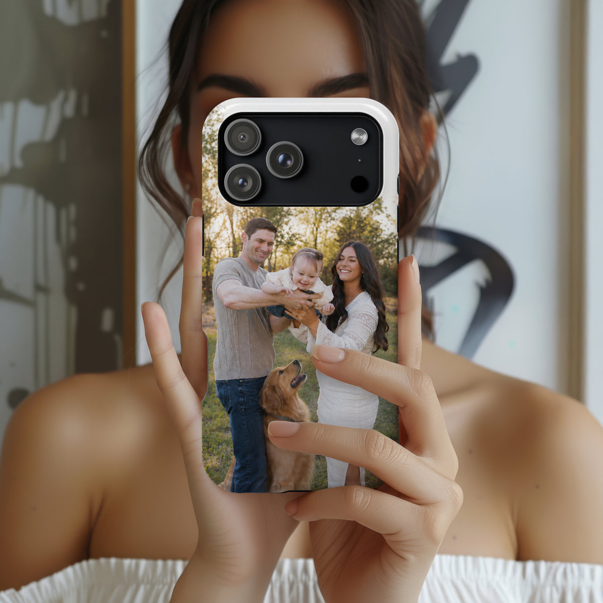 A person holds a smartphone in front of their face, showing a custom printed photo case on the phone. The case features a custom family photograph of a man, woman, and child with a golden retriever dog, displayed against a blurred indoor background.