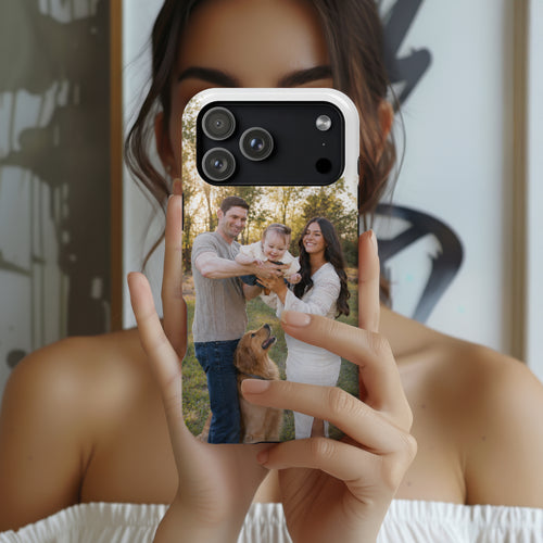 A person holds a smartphone in front of their face, showing a custom printed photo case on the phone. The case features a custom family photograph of a man, woman, and child with a golden retriever dog, displayed against a blurred indoor background.
