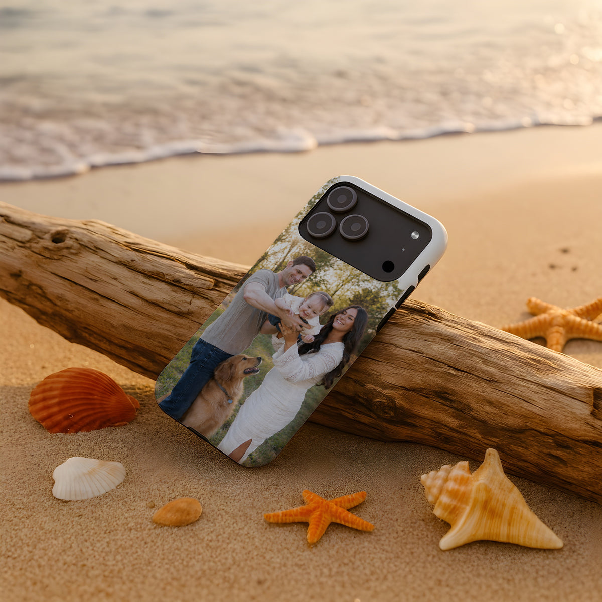 A custom smartphone case with a custom family photo print, shown resting on a beach sandlog with seashells and starfish. The case features a custom photo of a family of three and a golden retriever dog.