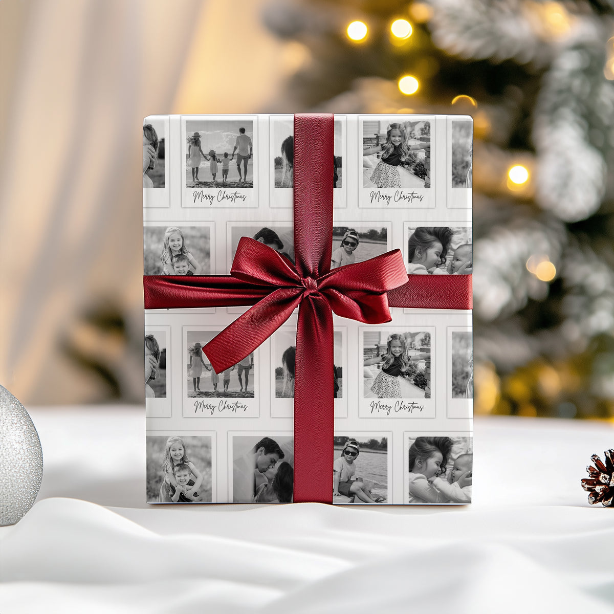 A wrapped gift box with a white holiday photo wrap featuring black and white family photos and 'Merry Christmas' text, tied with a wide red satin ribbon and bow, set on a white surface with a blurred Christmas tree in the background.