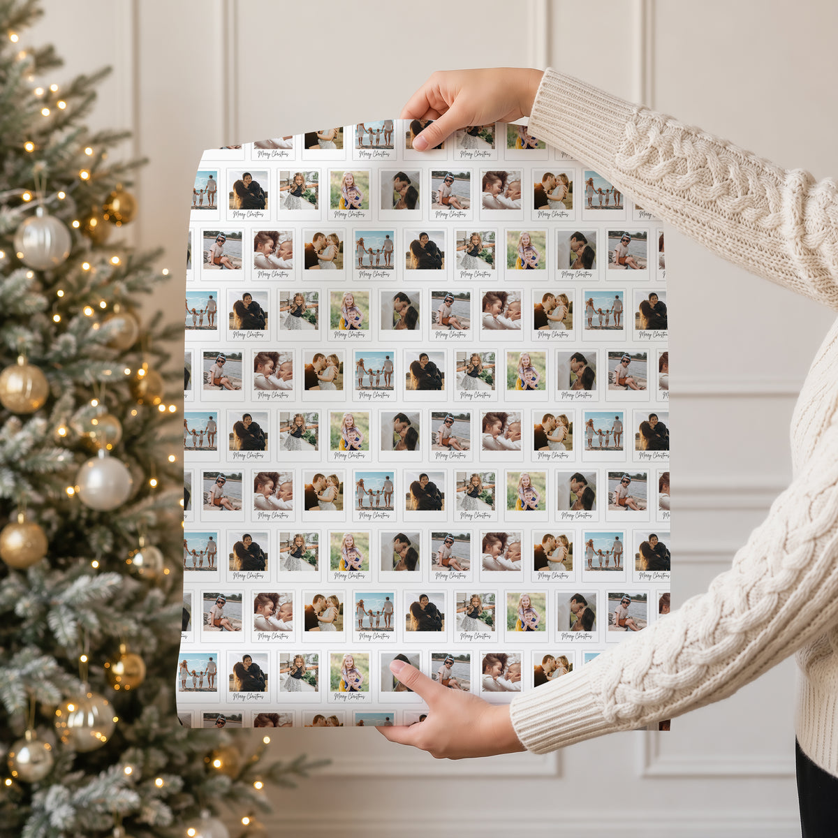 A person in a cream cable knit sweater holds up a large sheet of custom photo wrapping paper featuring a grid of small family photographs. The background shows a decorated Christmas tree with gold and silver ornaments on the left, and a white panelled wall on the right.
