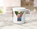 A white ceramic mug with a custom photo collage printed on the side, displayed on a light marble countertop with a blurred kitchen background.