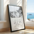 A framed custom baby nursery print leaning against a wall next to a window with a sea view. The black frame contains a black and white photo of a baby and printed text reading 'In this moment time stood still 'LUCAS JAMES BROWN' with birth details below.