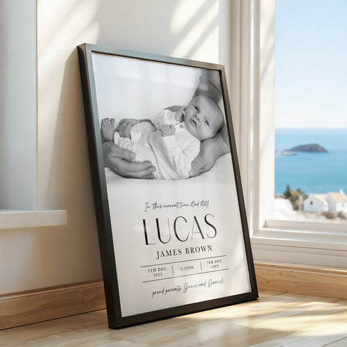 A framed custom baby nursery print leaning against a wall next to a window with a sea view. The black frame contains a black and white photo of a baby and printed text reading 'In this moment time stood still 'LUCAS JAMES BROWN' with birth details below.