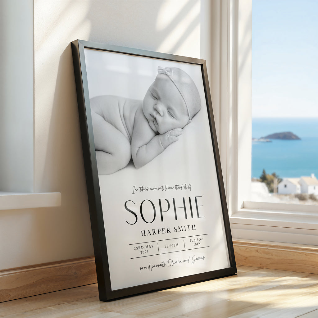 A framed personalised baby nursery print leaning against a window in a sunlit room. The print shows a black-and-white photo of a sleeping newborn baby with the text 'In this moment time stood still 'SOPHIE HARPER SMITH' and birth details.