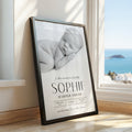 A framed personalised baby nursery print leaning against a window in a sunlit room. The print shows a black-and-white photo of a sleeping newborn baby with the text 'In this moment time stood still 'SOPHIE HARPER SMITH' and birth details.
