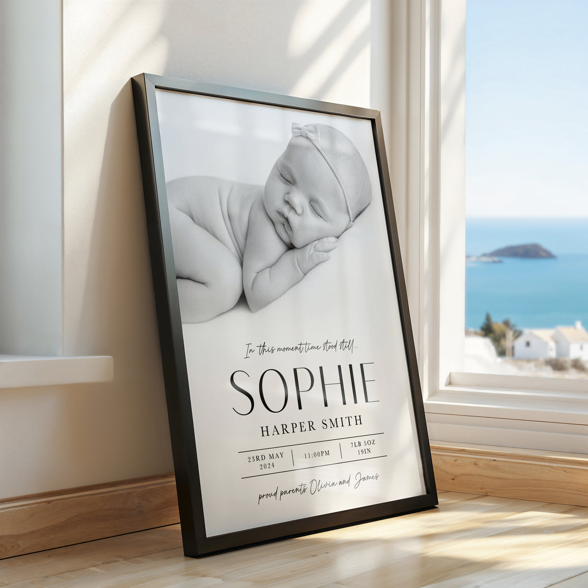 A framed personalised baby nursery print leaning against a window in a sunlit room. The print shows a black-and-white photo of a sleeping newborn baby with the text 'In this moment time stood still 'SOPHIE HARPER SMITH' and birth details.