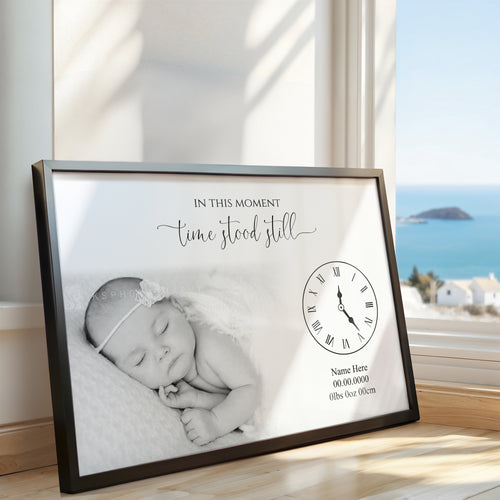 A black-framed personalized baby photo print leaning against a sunlit wall next to a window with an ocean view. The print features a black-and-white photo of a sleeping newborn, elegant script text reading 'IN THIS MOMENT time stood still,' a black clock face illustration, and placeholder text for the baby's name and birth details.