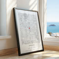 A framed personalised map print leaning against a window on a wooden floor, showing a black and white map with a small red heart and the text 'ENGAGED' and date/location details. The window reveals a bright blue ocean view.