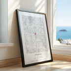 A framed personalised map print leaning against a window on a wooden floor, showing a black and white map with a small red heart and the text 'ENGAGED' and date/location details. The window reveals a bright blue ocean view.