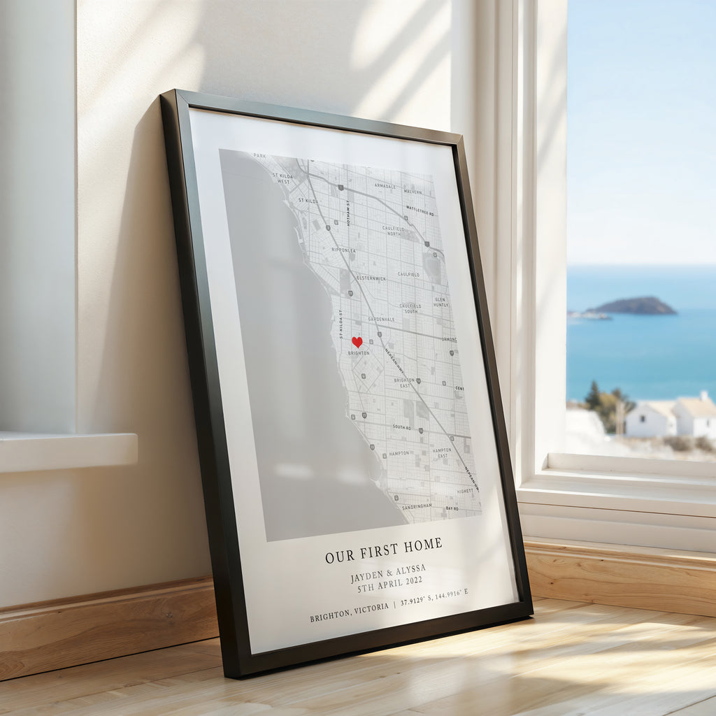 A framed personalised map print leaning against a wall on a wooden floor, with a bright coastal ocean view visible through the window behind it. The print has a black frame with a black and white map with a small red heart marker and the text 'OUR FIRST HOME' and custom names/date at the bottom.