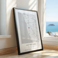 A framed personalised map print leaning against a wall on a wooden floor, with a bright coastal ocean view visible through the window behind it. The print has a black frame with a black and white map with a small red heart marker and the text 'OUR FIRST HOME' and custom names/date at the bottom.