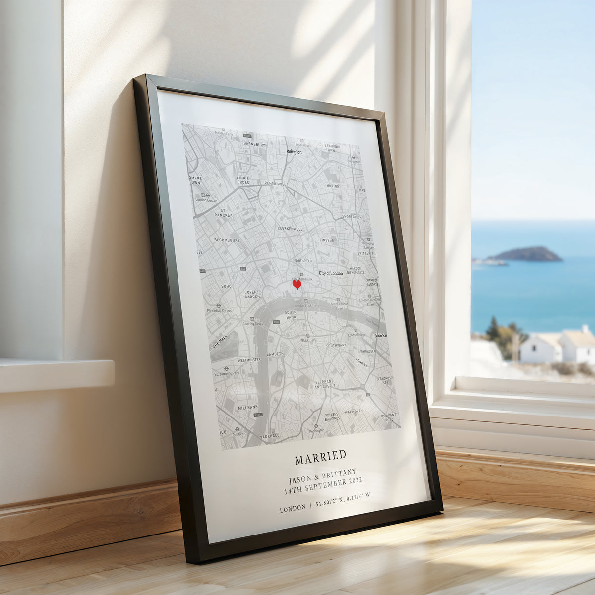 A framed custom map print leaning against a window on a wooden floor, showing a black and white street map with a small red heart marker and the text 'MARRIED' and names below. The window reveals a bright blue sea and distant island.