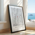A framed custom map print leaning against a wall on a wooden floor, with a window and ocean view in the background. The print features a black and white map of New York City with a small red heart marking and the text 'WHERE IT ALL BEGAN' with personalised names and date.