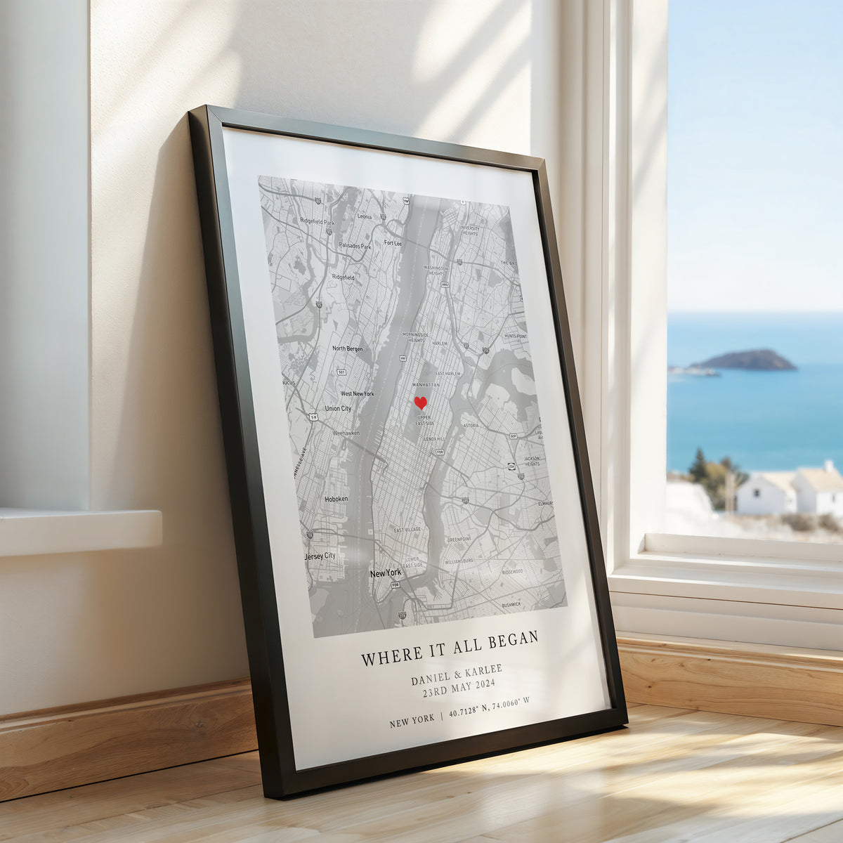 A framed custom map print leaning against a wall on a wooden floor, with a window and ocean view in the background. The print features a black and white map of New York City with a small red heart marking and the text 'WHERE IT ALL BEGAN' with personalised names and date.