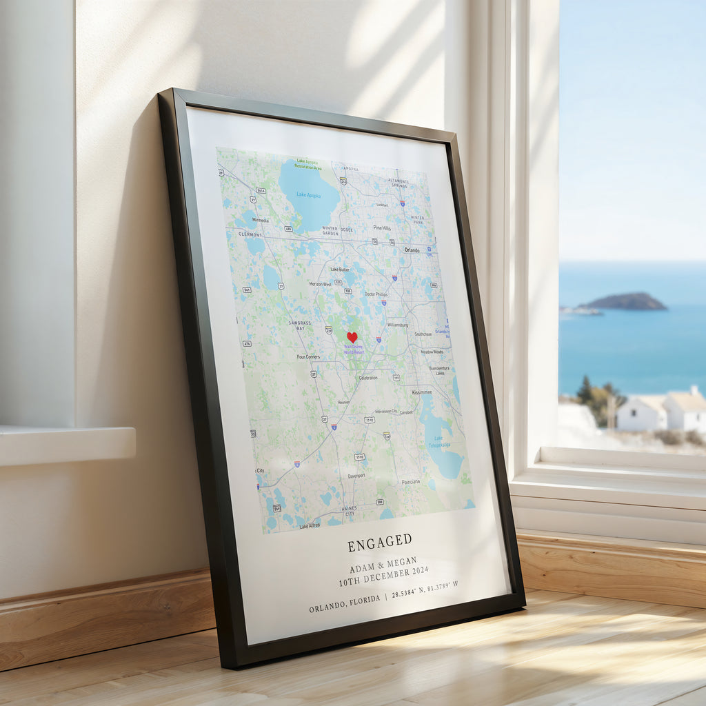 A framed personalised map print leaning against a window on a wooden floor, with a coastal ocean view outside. The print has a black frame and a map with a small red heart near the centre and text at the bottom reading 'ENGAGED'.