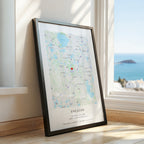 A framed personalised map print leaning against a window on a wooden floor, with a coastal ocean view outside. The print has a black frame and a map with a small red heart near the centre and text at the bottom reading 'ENGAGED'.