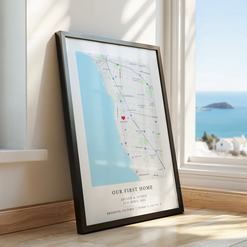 A framed personalised map print leaning against a window on a wooden floor, showing a coastal street layout with the text 'OUR FIRST HOME' and names/date/location. The room has warm cream walls, natural sunlight, and a blue ocean view outside.