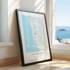 A framed personalised map print leaning against a window on a wooden floor, showing a coastal street layout with the text 'OUR FIRST HOME' and names/date/location. The room has warm cream walls, natural sunlight, and a blue ocean view outside.