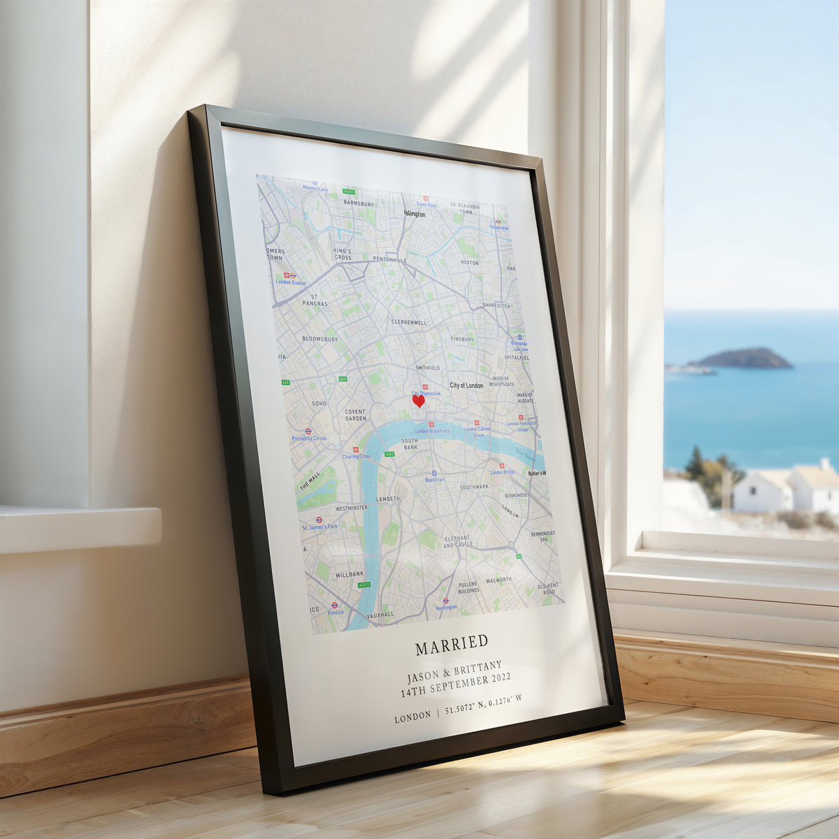 A framed custom map print leaning against a wall next to a window with a coastal view. The black framed print features a detailed map of London with a red heart marker and the text 'MARRIED' and names/date printed at the bottom.