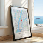 A framed New York City map print leaning against a window on a wooden floor, with a bright ocean view outside. The print has a black frame and the text 'WHERE IT ALL BEGAN' with names and date printed below the map.