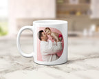 A white ceramic coffee mug with a custom printed photo of a father holding and kissing his baby, displayed on a light marble countertop with a blurred kitchen background.