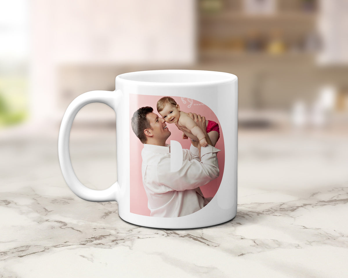 A white ceramic coffee mug with a custom printed photo of a father holding and kissing his baby, displayed on a light marble countertop with a blurred kitchen background.