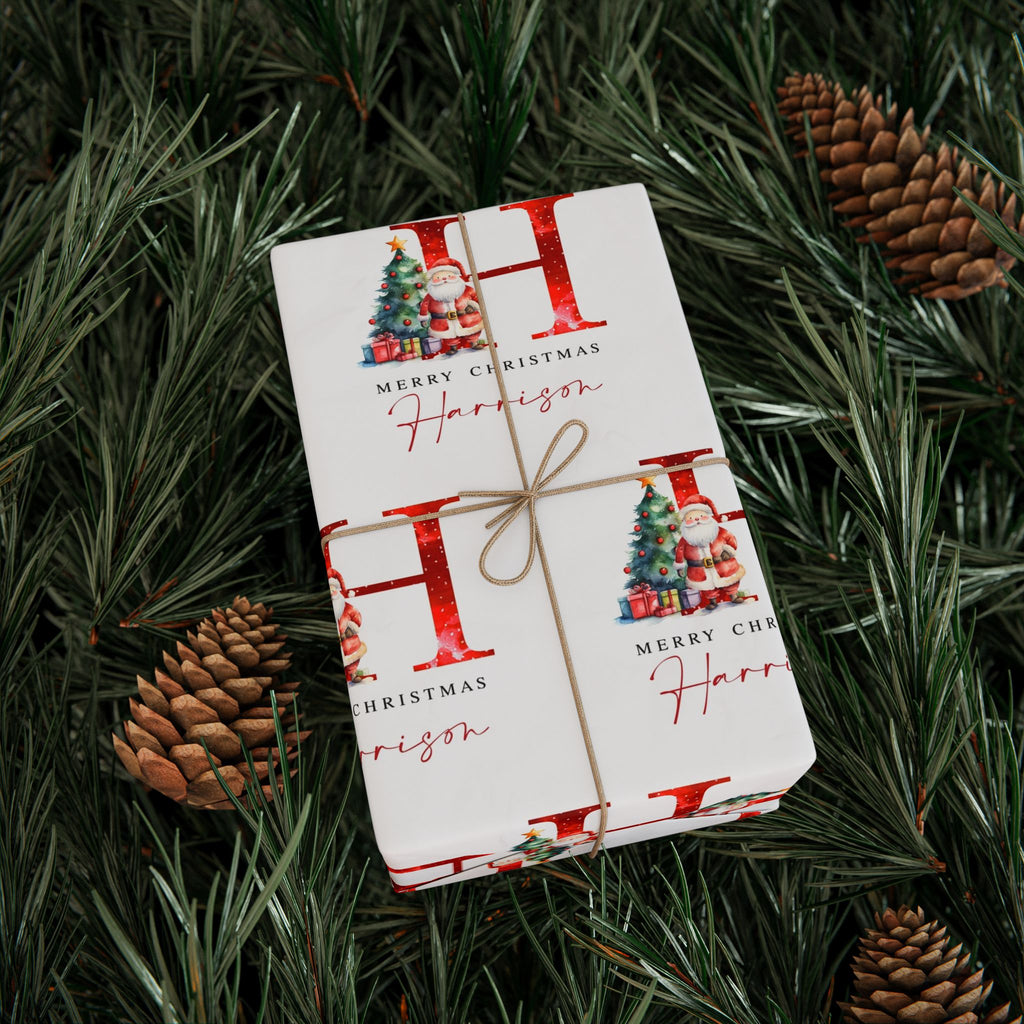 A wrapped Christmas gift box tied with twine, placed on green pine branches with pinecones. The wrapping paper is white with large red letters, small Christmas tree illustrations, and the text 'Merry Christmas' and 'Harrison'.