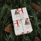 A wrapped Christmas gift box tied with twine, placed on green pine branches with pinecones. The wrapping paper is white with large red letters, small Christmas tree illustrations, and the text 'Merry Christmas' and 'Harrison'.