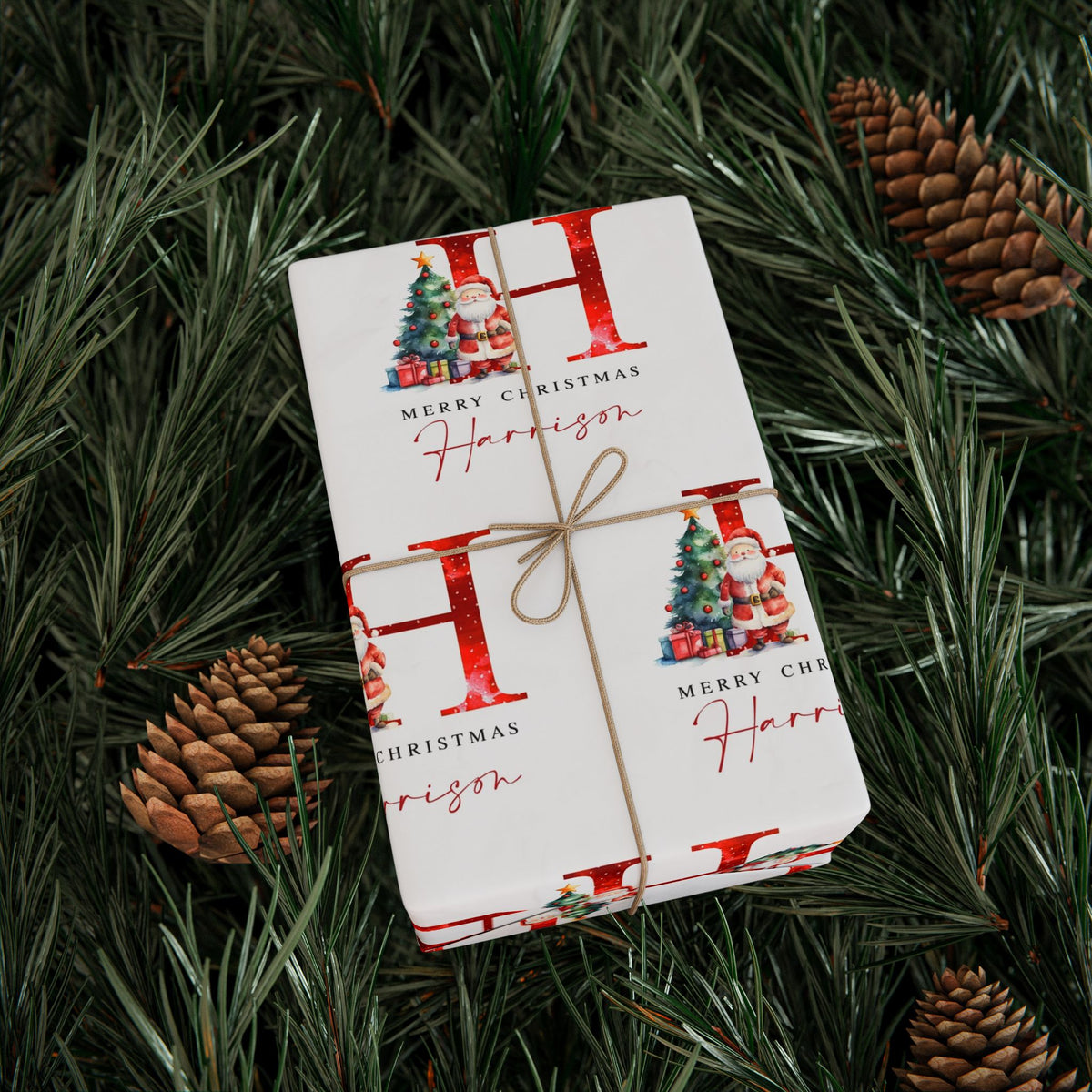 A wrapped Christmas gift box tied with twine, placed on green pine branches with pinecones. The wrapping paper is white with large red letters, small Christmas tree illustrations, and the text 'Merry Christmas' and 'Harrison'.