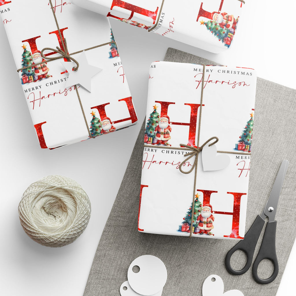 Three wrapped Christmas gift boxes on a light gray surface with a gray cloth, wrapped in white paper printed with large red letters, 'Merry Christmas' text, Santa illustrations, and a twine bow. A ball of twine, scissors, and round tags are arranged nearby.