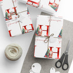 Three wrapped Christmas gift boxes on a light gray surface with a gray cloth, wrapped in white paper printed with large red letters, 'Merry Christmas' text, Santa illustrations, and a twine bow. A ball of twine, scissors, and round tags are arranged nearby.