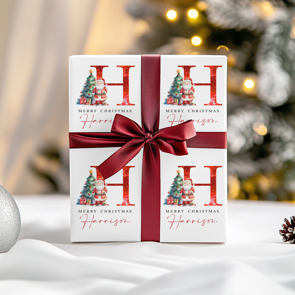 A personalised Christmas gift box wrapped in a dark red satin ribbon and bow, featuring a white printed design with a large red 'H', 'Merry Christmas', and the name 'Harrison' in cursive. The background is a softly blurred festive Christmas tree with warm lights.