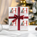 A personalised Christmas gift box wrapped in a dark red satin ribbon and bow, featuring a white printed design with a large red 'H', 'Merry Christmas', and the name 'Harrison' in cursive. The background is a softly blurred festive Christmas tree with warm lights.