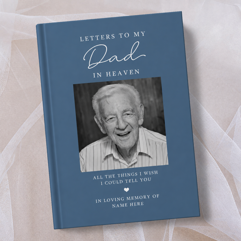 A personalised hardcover book titled 'Letters to My Dad in Heaven' with a slate blue cover, a black and white portrait photo of an elderly man, and white text, displayed on a soft beige fabric background.