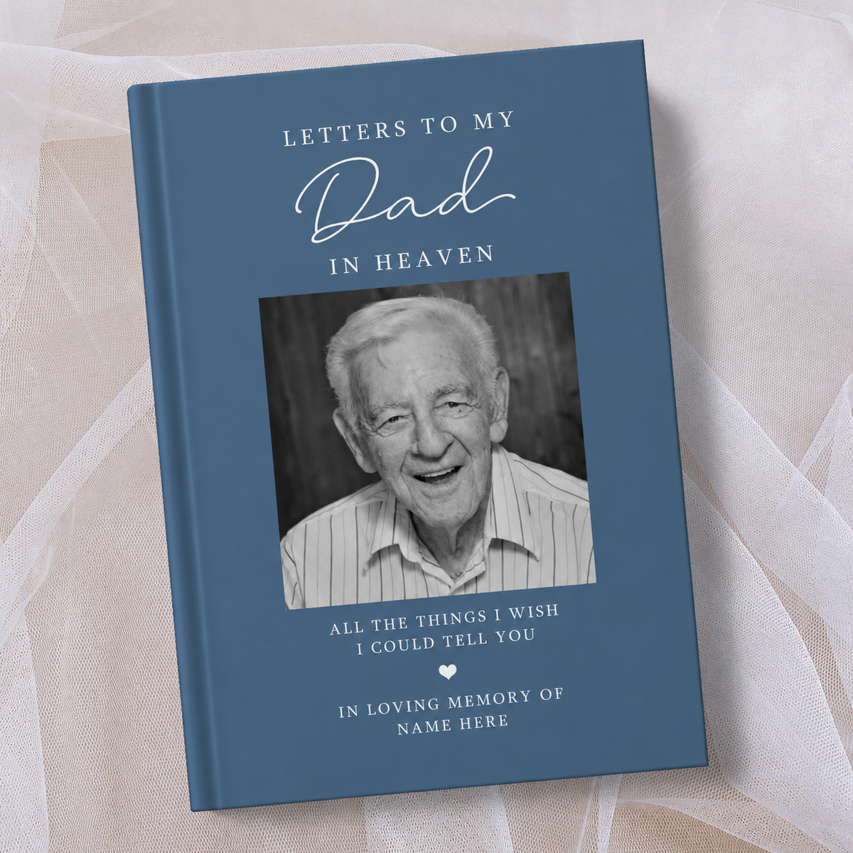 A personalised hardcover book titled 'Letters to My Dad in Heaven' with a slate blue cover, a black and white portrait photo of an elderly man, and white text, displayed on a soft beige fabric background.