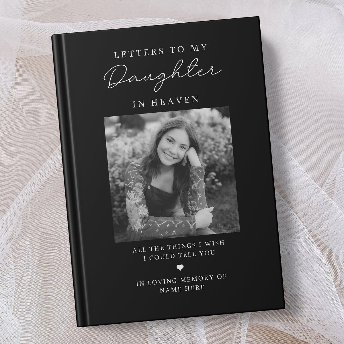 A hardcover memorial book titled 'Letters to My Daughter in Heaven' with a black cover, white script and serif text, and a placeholder black and white photo of a young woman. The book is photographed on soft white draped fabric.