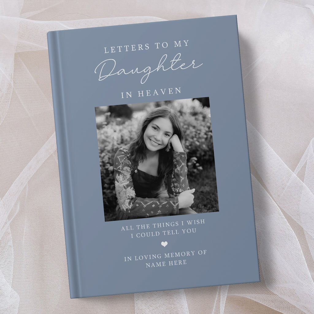 A personalised hardcover book titled 'Letters to My Daughter in Heaven' in a muted slate blue colour, featuring a customisable black and white photo of a young woman on the front cover, displayed on a soft beige fabric background.