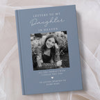 A personalised hardcover book titled 'Letters to My Daughter in Heaven' in a muted slate blue colour, featuring a customisable black and white photo of a young woman on the front cover, displayed on a soft beige fabric background.