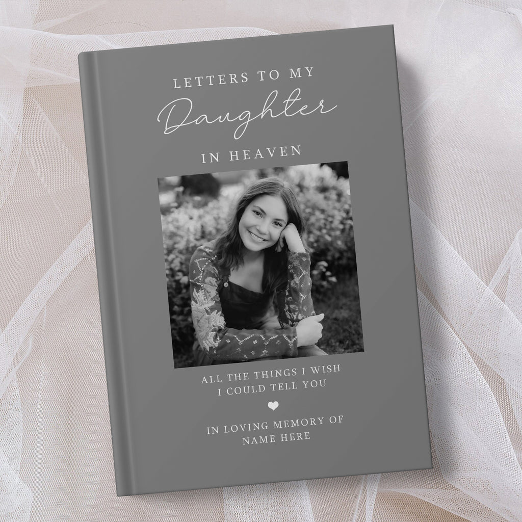 A personalised hardcover book titled 'Letters to My Daughter in Heaven' with a grey cover, featuring a placeholder black and white photo of a young woman and memorial text, styled on a soft beige fabric background.