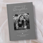 A personalised hardcover book titled 'Letters to My Daughter in Heaven' with a grey cover, featuring a placeholder black and white photo of a young woman and memorial text, styled on a soft beige fabric background.