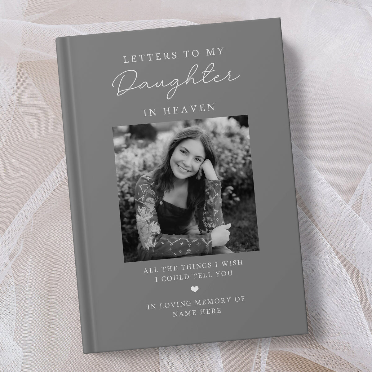 A personalised hardcover book titled 'Letters to My Daughter in Heaven' with a grey cover, featuring a placeholder black and white photo of a young woman and memorial text, styled on a soft beige fabric background.